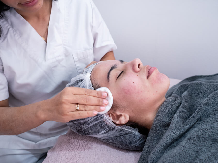 Beneficios del hydrafacial que-es - Profesional limpiando el rostro de una mujer recostada con gorro desechable