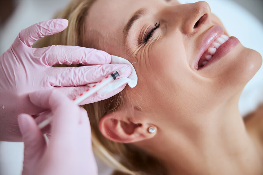 Nctf 135 ha antes y después opiniones - Mujer sonriendo mientras recibe una inyección estética cerca del ojo por un especialista con guantes rosados.