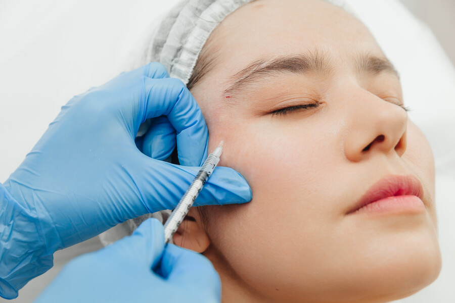 Bótox para la cara cuidados - Mujer recibiendo inyección estética en la zona del pómulo por profesional con guantes azules.