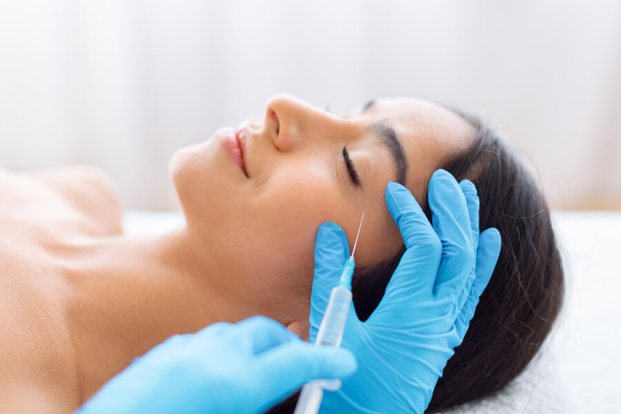 Bótox para la cara el procedimiento - Mujer recostada recibiendo una inyección estética en el rostro por profesional con guantes azules.