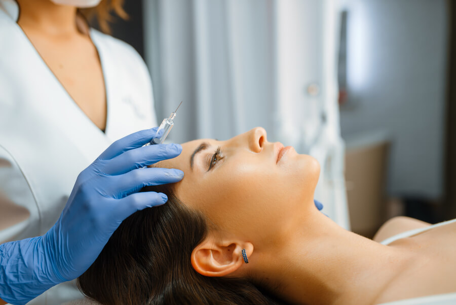 Harmonyca efectos secundarios conclusión - Mujer recostada recibiendo inyección estética en la frente por profesional con guantes.
