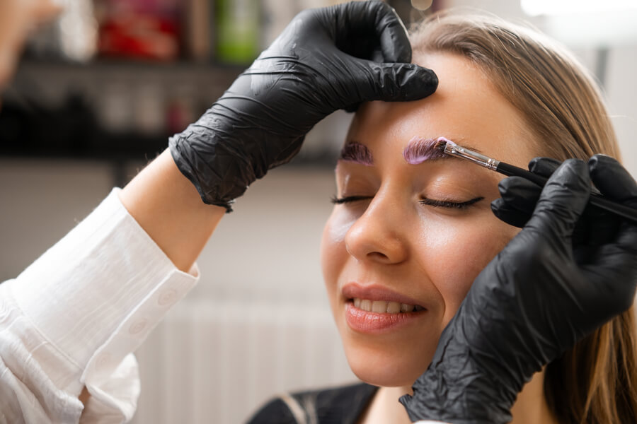 Cuanto dura la micropigmentación de cejas cuidados - Mujer con los ojos cerrados mientras un especialista aplica tinte en sus cejas con un pincel.